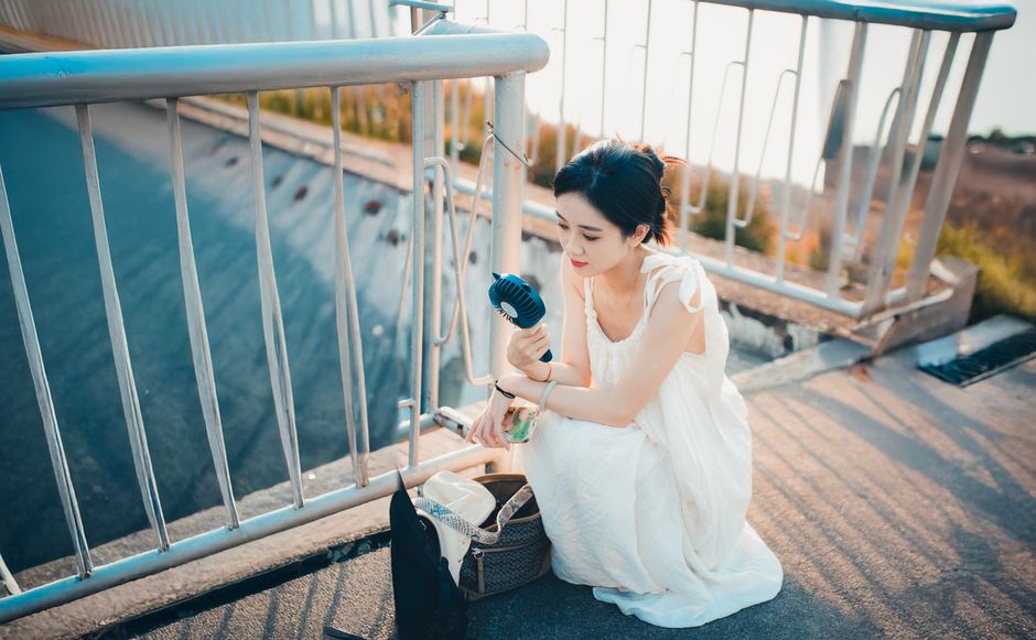晴れた日の屋外で、白いドレスを着た女性が携帯扇風機で涼を取っています。
