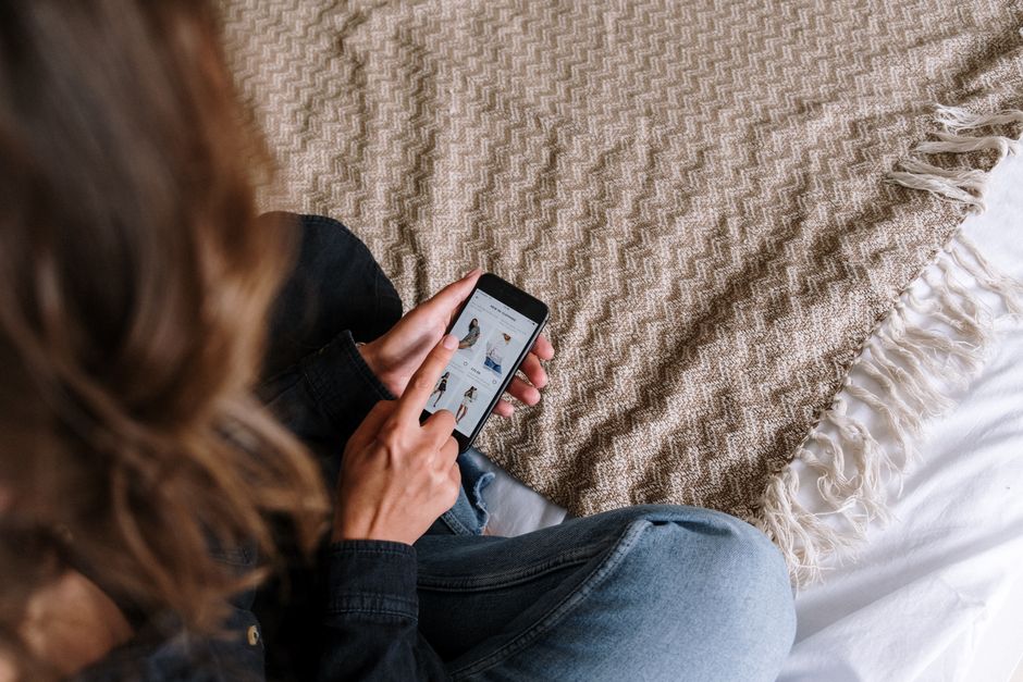女性がベッドに座って心地よいブランケットに包まれながら、スマートフォンでオンラインショッピングアプリを閲覧しています。