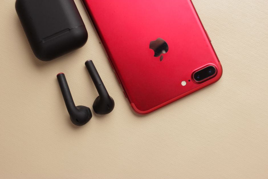 ベージュの表面で、黒いワイヤレスイヤフォンと充電ケースが置かれたスリムな赤いスマートフォンのクローズアップ。